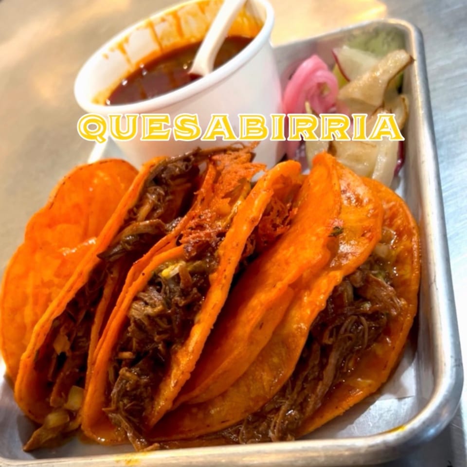 Four quesabirria tacos with a side of consommé and pickled onions on a metal tray.