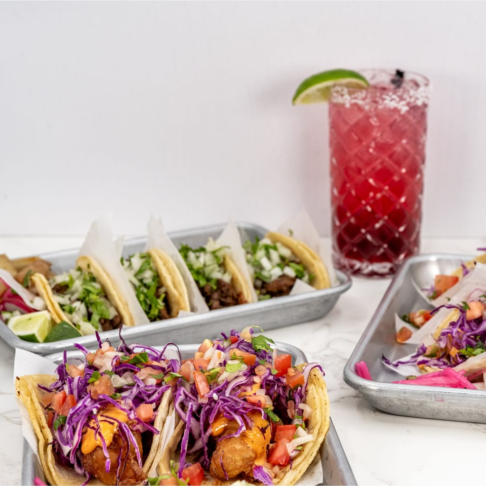 Assortment of Taco el Diablo tacos with various toppings and a red margarita on a white marble surface.