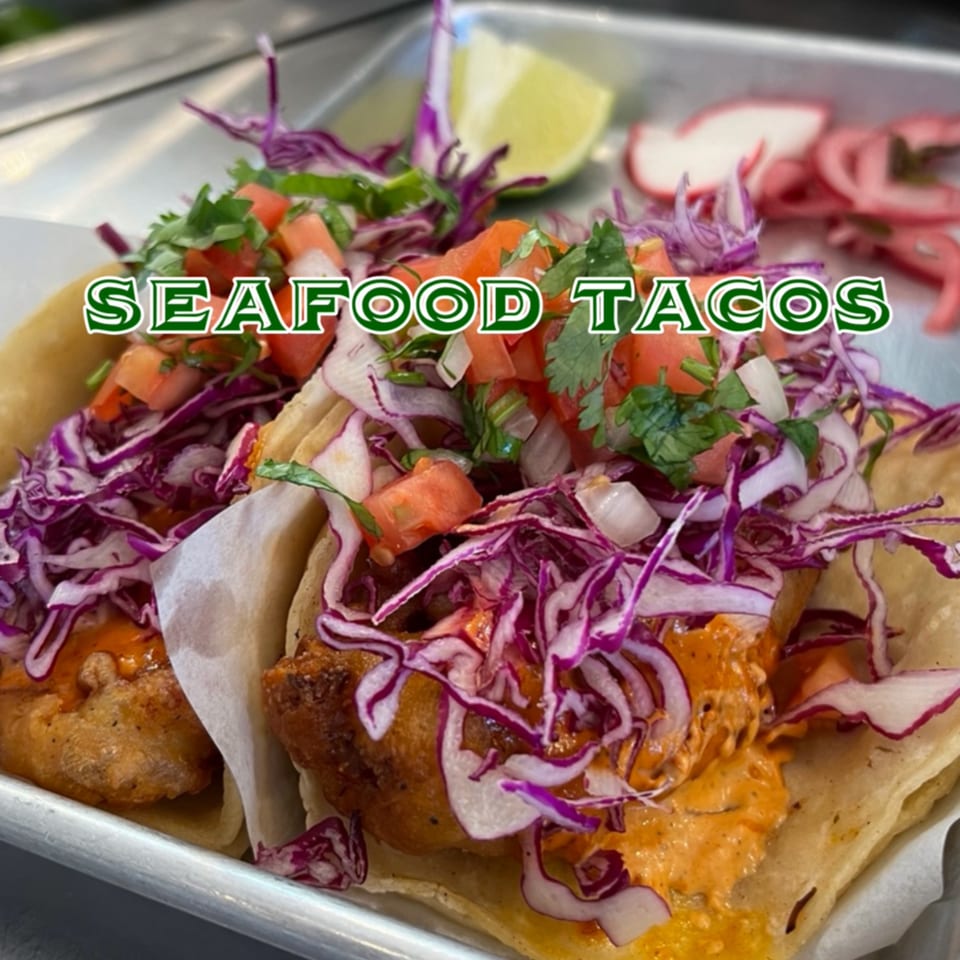 Two vibrant seafood tacos with fried fish, purple cabbage, pico de gallo, and orange sauce.