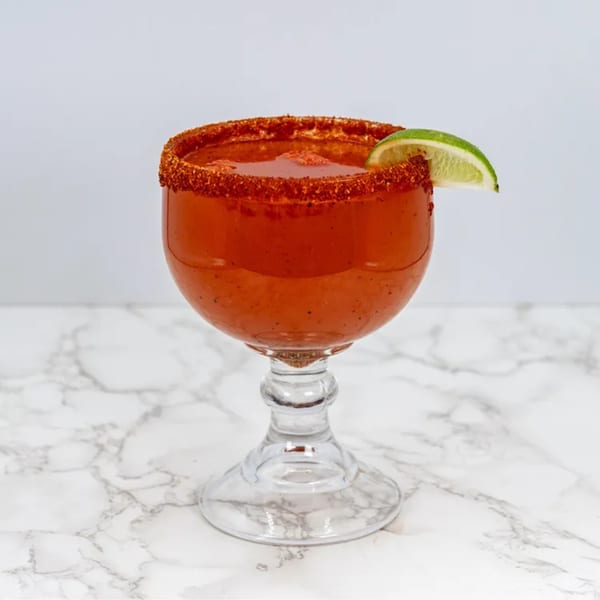 A red michelada cocktail in a goblet, rimmed with chili salt and garnished with a lime wedge.