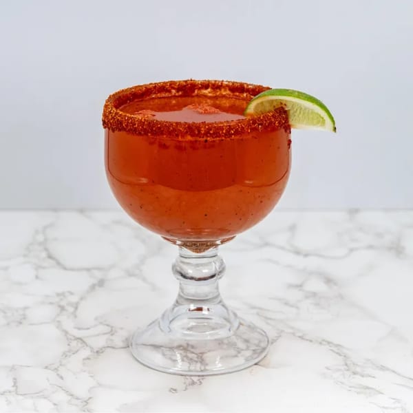 A red michelada cocktail in a goblet, rimmed with chili powder and garnished with a lime wedge.