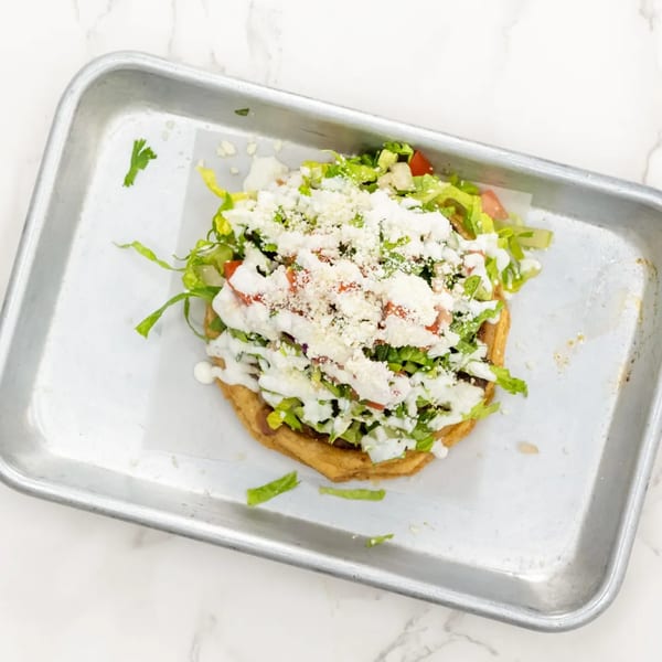 A Mexican tostada with lettuce, tomatoes, crema, and cotija cheese on a metal tray.