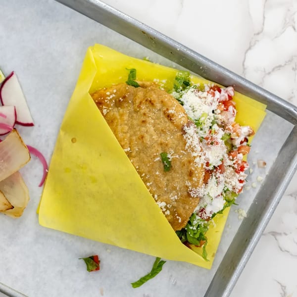 A golden-brown Mexican gordita, filled with lettuce, pico de gallo, and cotija cheese, wrapped in yellow paper.