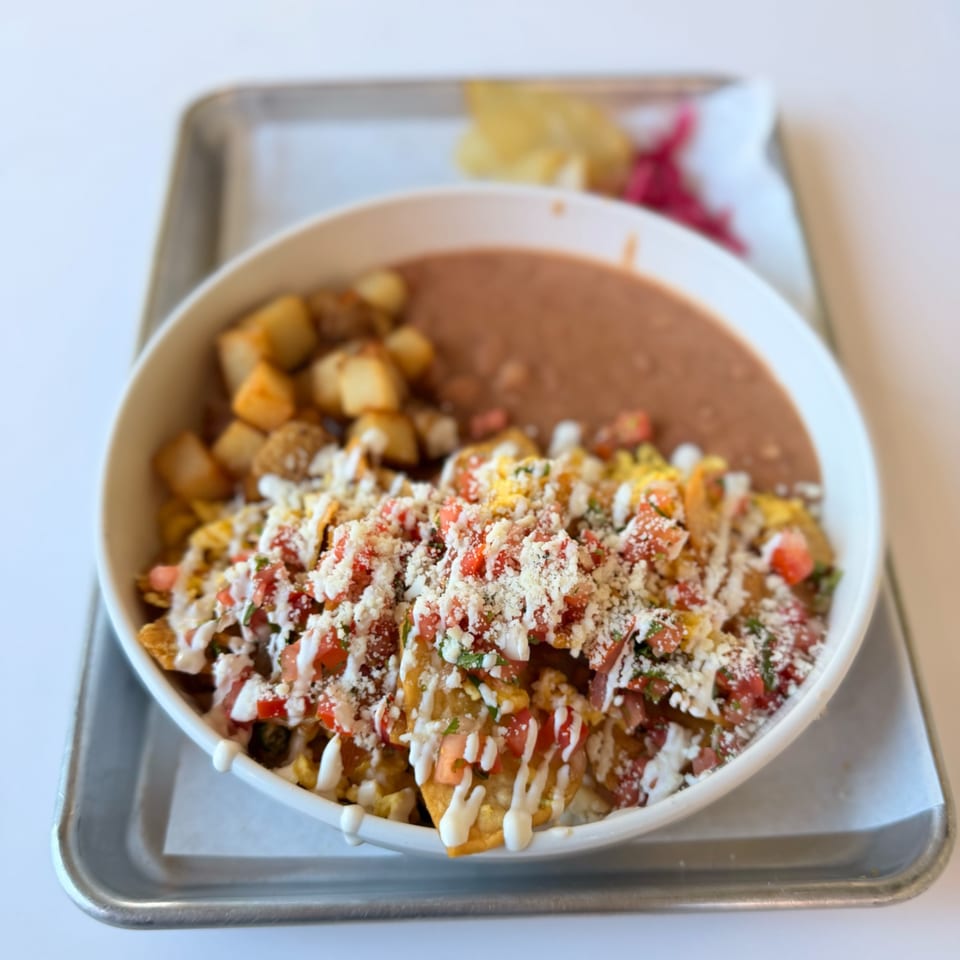 A bowl of Mexican chilaquiles with refried beans and potatoes on a metal tray.