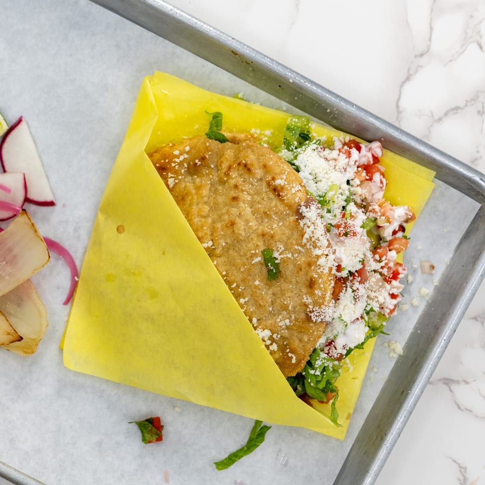 A golden-fried taco from Taco el Diablo, filled with lettuce, tomatoes, and cheese, wrapped in yellow paper.
