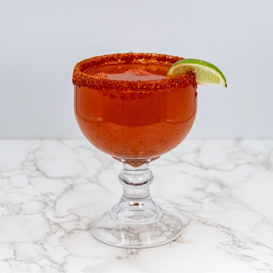 A red michelada cocktail in a goblet, rimmed with chili powder and garnished with a lime wedge.