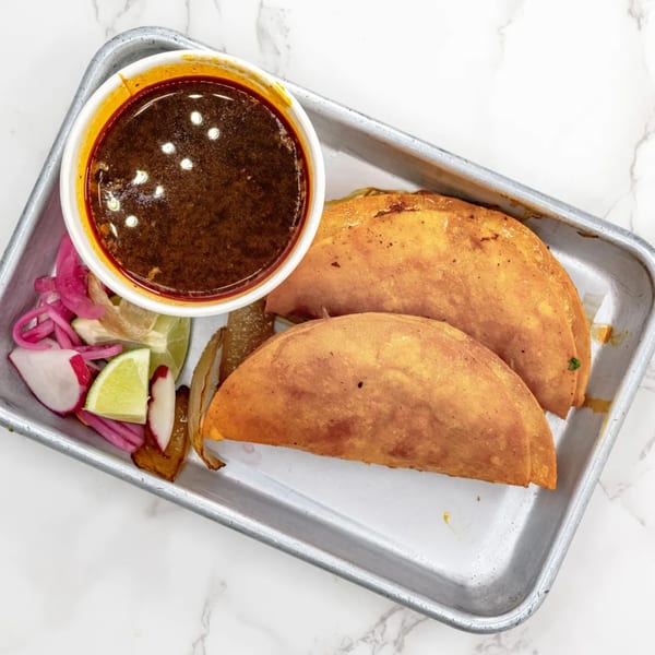 Two golden-brown birria tacos with a side of rich consommé, pickled onions, radishes, and lime wedges.