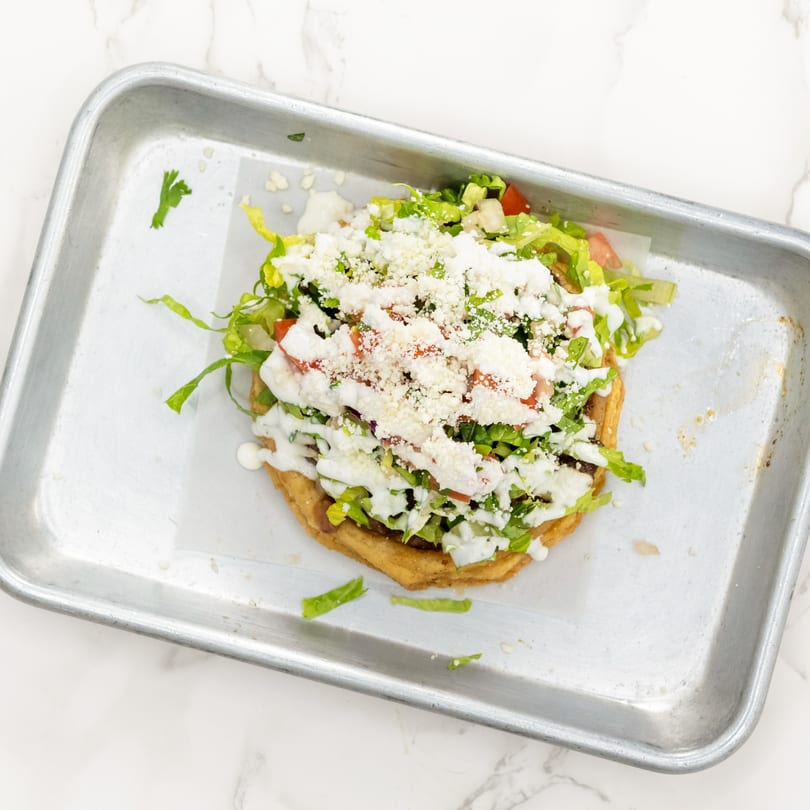 A Mexican sope with lettuce, tomatoes, sour cream, and cotija cheese on a metal tray.
