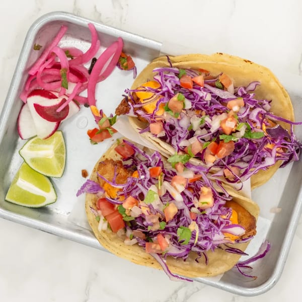 Two fish tacos with purple cabbage, pico de gallo, and sauce, served with pickled onions and lime wedges.