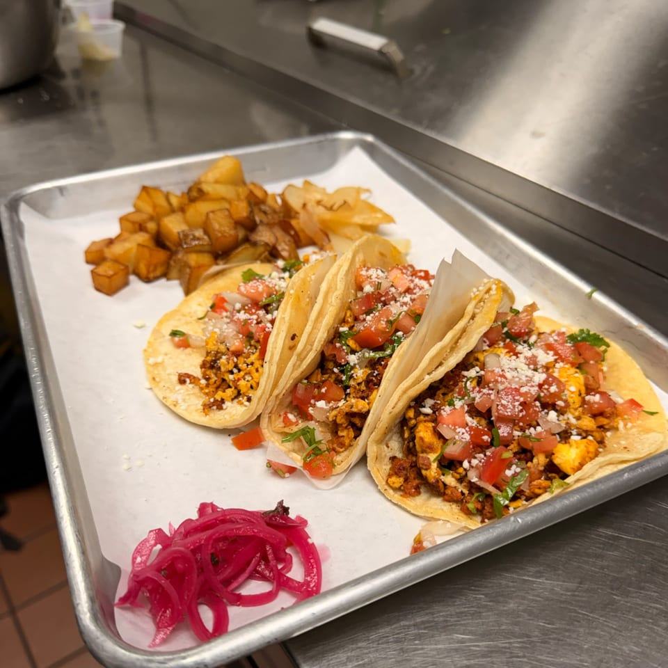 Three chorizo and egg breakfast tacos with diced tomatoes, cheese, and cilantro, served with potatoes and pickled onions.