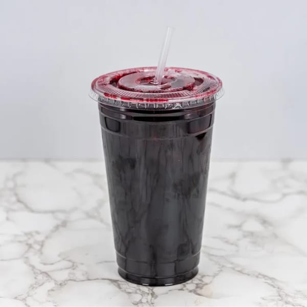 A dark red Mexican drink in a clear plastic cup with a lid and straw on a marble surface.