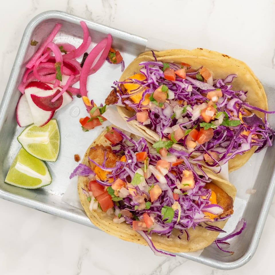 Two fish tacos with purple cabbage, pico de gallo, and sauce, served with pickled onions and lime wedges.
